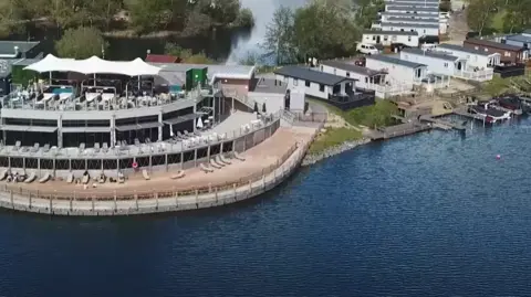 Away Resorts An aerial image of Tattershall Lakes Country Park shows a lakefront with several long, thin static caravans on the shore, and a couple of small boats moored up. There's also a large communal area and beach with sunloungers set out and a large white parasol offering some shade.