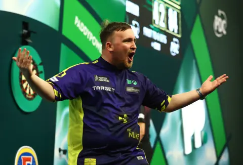 Reuters Luke Littler, a young man in a navy and yellow sports top, shouts at the crowd on a stage. There is a darts board behind him