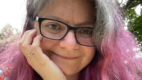 Dimitra Fimi Dimita Fimi, who has wavy grey and dyed-pink hair and is wearing glasses, smiling and looking at the camera with her chin propped on her hand