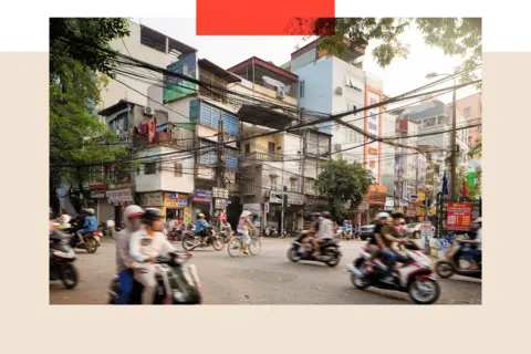 Getty Images Morning traffic on Lo Duc Street in Hanoi, Vietnam on a warm spring day. People are commuting on bikes and motorbikes, or walking and shopping. Apartment buildings are rising above the street behind electric cables.
