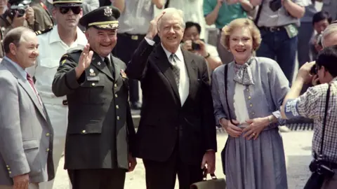 AFP Former U.S. President Jimmy Carter and his wife Rosalynn wave to journalists, surrounded by a crowd. He wears a dark suit, and she wears blue skirt, jacket and scarf