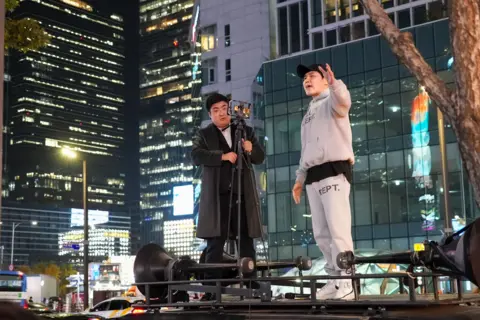 A man in a grey hooded top and black cap is pictured shouting on top of a van with speakers as he is live streamed 