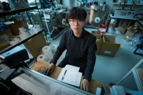 A man wearing glasses and a dark coloured shirt sits at a desk, writing notes in a piece of paper and using a computer 