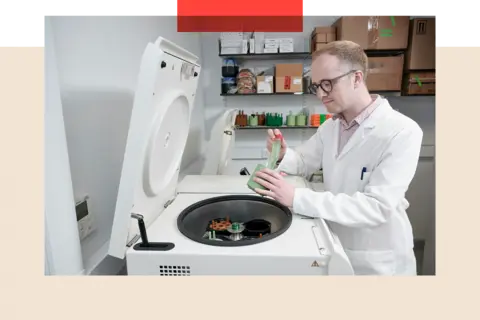 A white machine with a circular hatch contains scientific equipment. A man in a white coat is leaning over the hatch and working with some of the equipement