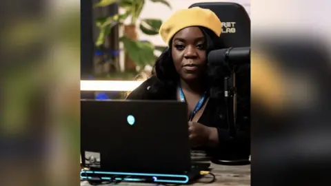 Jonas Kontautas A woman in a yellow beret sits at a desk in a high-backed gaming chair. A streaming microphone is set up close to her face and there's a black laptop with a glowing alien logo in the middle of the lid open in front of her.