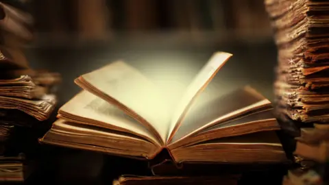 Getty Images Two stacks of papers with an open book lying between them on a desk and a bright light shining from within the pages