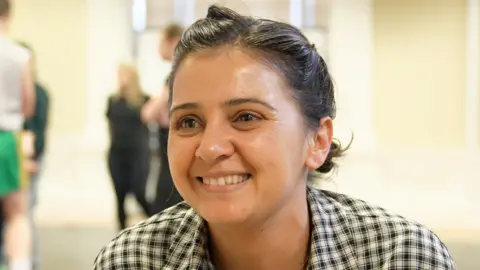 BBC A smiling Natalie Davies, a Bradford-born actor and writer. She is wearing a black and white checked shirt and has her black hair tied back.