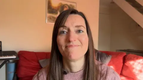 Georgia Poncia/BBC Natasha has long brown hair and is wearing a lilac top and sitting on a red sofa. 