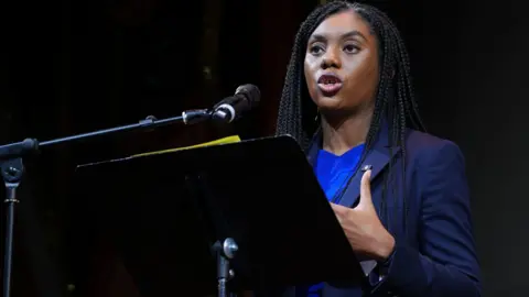 PA Media Conservative leader Kemi Badenoch, standing behind a black lectern on a stage, speaks to an audience  