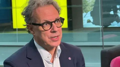 BBC A man wearing a dark suit and glasses looks to the right of the camera while he talks