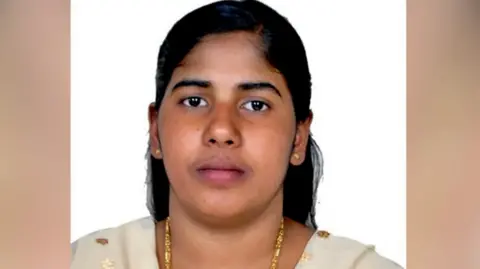 BBC A picture of Nimisha Priya wearing gold jewellery, her hair tied back. She is wearing a beige-coloured Indian suit. 