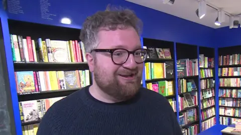 BBC A medium close up of a smiling Tom Rowley stood in his shop bookshop in south London with books on blue shelves in background.